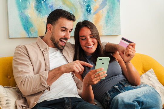 A joyful couple uses a smartphone while sitting close on a yellow sofa, one points at the device as they browse and consider a purchase, exuding excitement and connection