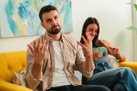 A candid moment captures a man gesturing with open hands while seated on a bright yellow sofa while a woman nearby focuses on a smartphone creating a relaxed and connected mood in a cozy living space.