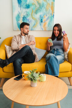 Two people sit close on a bright yellow couch smiling and chatting while one checks a phone and the other holds a card on a tidy coffee table. The moment highlights connection and everyday tech use up