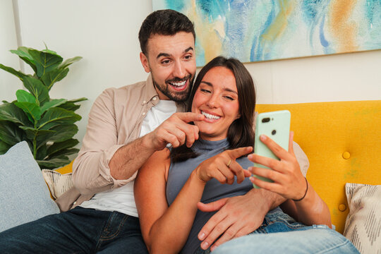 A cheerful couple sits close on a soft yellow couch, capturing a spontaneous selfie with a light green smartphone. They smile broadly, point toward the screen, and share a joyful moment of connection
