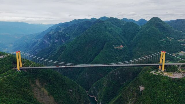 Aerial pull back of a huge suspension bridge spanning a deep mountain canyon under an overcast sky. Dramatic engineering infrastructure landscape.