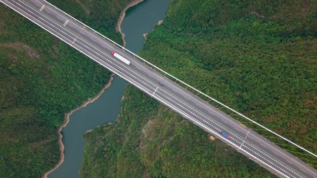 Aerial hover shot of modern suspension bridge and highway spanning across deep green valley and reservoir on overcast day.