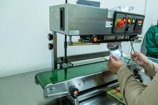 Worker hands operating industrial packaging machine sealing aluminum foil pouch in clean production environment.