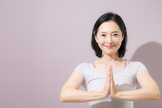 A middle-aged Japanese woman performing a yoga pose with her hands pressed together in front of her chest, looking at the camera as she relaxes and brings her mind and body into balance