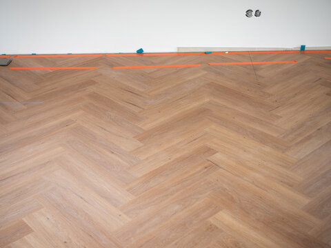 Light oak look vinyl flooring in herringbone pattern with spacers and installation markers along the wall, showing finishing work before baseboard installation during renovation.