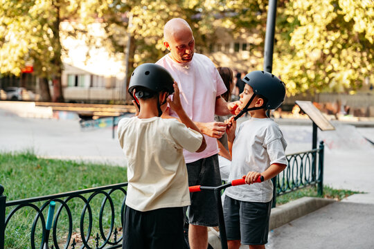 Two children learn to ride scooters at a skate park with an adult in the afternoon light