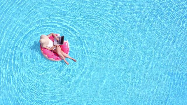 Top down view of woman working on laptop in swimming pool, aerial drone shot of freelance digital nomad on pink donut inflatable float, creative remote business executive summer lifestyle vacation