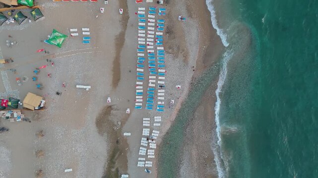 An aerial overhead drone shot moving upward over a beach resort, revealing rows of sun loungers, resort buildings, and waves crashing on the shore.