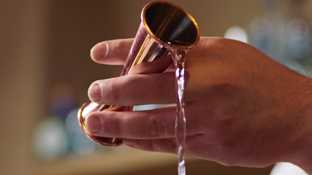 Close up shot of a professional bartender's hand using a stainless steel measuring jigger to pour a perfect shot of tequila into a glass at a bar