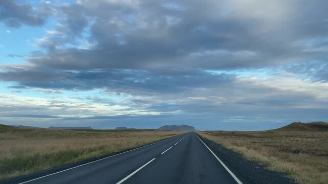 Driver point of view traveling on asphalt highway through the open Icelandic highlands beneath expansive clouds and distant mountains.