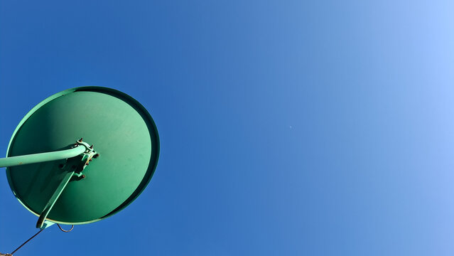 Minimalist bottom view of a green parabolic satellite receiver dish isolated on a vast clear blue sky. Symbol for satellite communication, media transmission, and signal network.