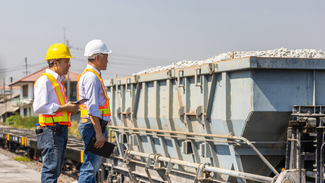 Railway engineers with tablet and safety equipment checking railway transport and logistics at cargo yard, industrial workers in safety vests and hard hats inspecting freight train and cargo
