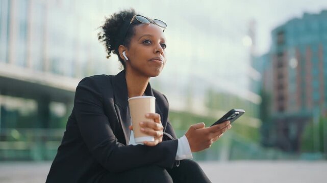 Self-confident black woman enjoying hot coffee and favorite music in earphones. Portrait of successful businesswoman or executive in downtown in morning, energy for new working day, relax after work