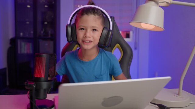 A young content creator wearing headphones talks to the camera in front of a laptop and microphone, then raises his hand to wave goodbye in a colorfully lit home studio.