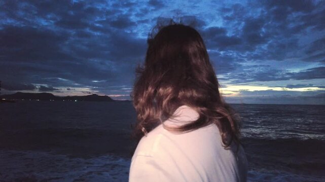 A man with windblown hair stands facing the sea at dusk while waves roll in and clouds drift across the darkening sky.