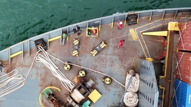 multiple ship crew members in bright safety orange working with massive ropes and heavy machinery on the vibrant green deck of a large vessel's bow, with blue water surrounding