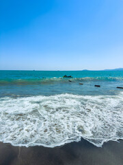 Waves crash on the shore as boats drift in the distance under the clear blue sky © DSMT