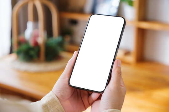 Mockup image of a woman holding and using mobile phone with blank desktop white screen at home