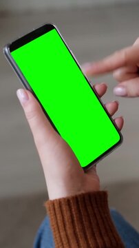 Female hands scrolling and swiping on a clean green screen chroma key smartphone mockup template indoor. Close up of woman finger touching mobile phone touchscreen blank display. Vertical video