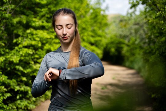 Sporty woman monitoring her fitness activity on a smartwatch during a run through a sunny green forest.