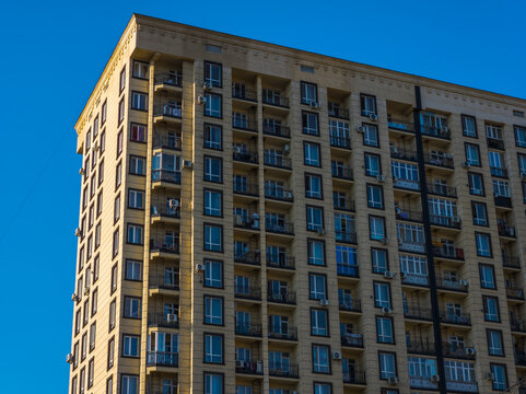 Detailed view of a tall apartment building with a light yellow facade. Numerous windows and balconies with black railings are visible, illuminated by sunlight against a clear blue sky.