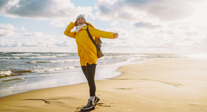 Traveler with backpack walks along sandy beach at sunset. Woman wearing bright raincoat and carrying backpack feels sense of freedom, enjoys seascape. Concept of adventure, freedom, and enjoyment.
