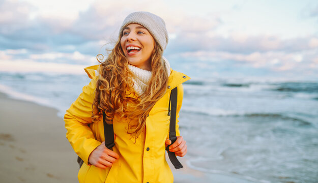 Traveler with backpack walks along sandy beach at sunset. Woman wearing bright raincoat and carrying backpack feels sense of freedom, enjoys seascape. Concept of adventure, freedom, and enjoyment.