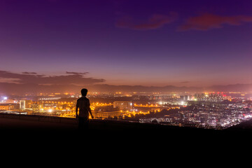 A silhouetted person stands at a high overlook, gazing at a brightly lit cityscape under a purple dusk sky. © tai