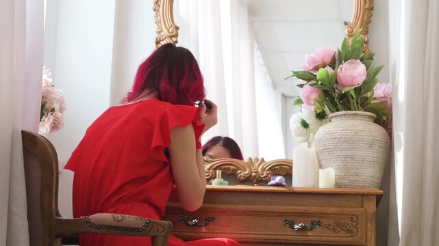 Young Woman with Red Hair in a Red Dress Applying Makeup in Front of a Vintage Vanity Mirror