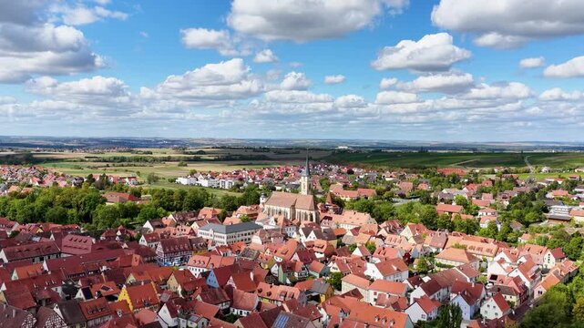 Malerische Panorama-Aussicht auf die historische Altstadt von Iphofen mit dem markanten Stadtturm, umgeben von gr&uuml;nen B&auml;umen unter einem blau-wei&szlig;en Sommerhimmel.