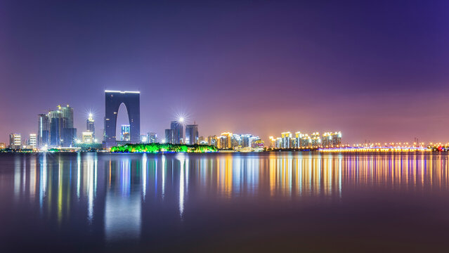 A vibrant night city skyline with illuminated buildings and their colorful reflections on calm water under a purple-hued sky.