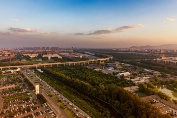 Fototapeta premium Aerial panorama of urban area featuring elevated overpass, roads, green spaces and buildings under sunset sky.
