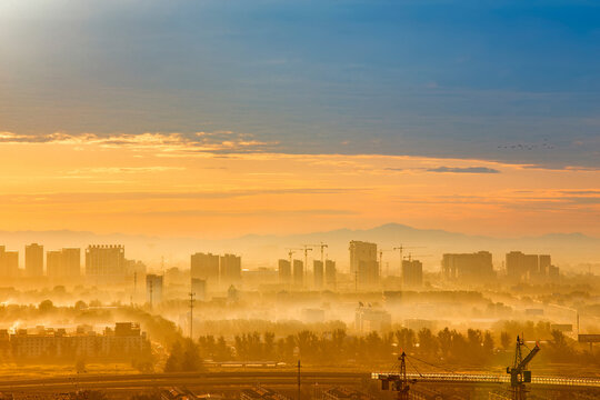 A misty urban landscape shows a distant city skyline under warm sunrise light, with construction cranes visible.