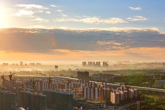A hazy urban cityscape at sunrise shows ongoing construction with cranes and distant buildings under a colorful sky.