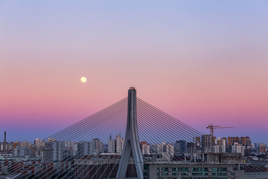 A cable-stayed bridge stands against a dusk sky with a bright moon above a city skyline with construction cranes.