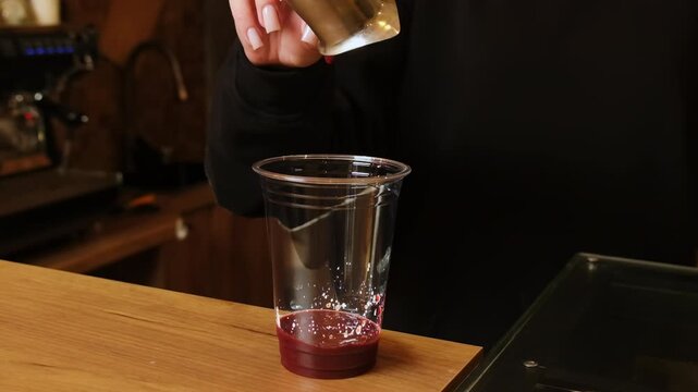 Barista drains last cherry jam into clear plastic cup. Woman tilts metal measure letting single red drop fall onto sweet fruit base slowly