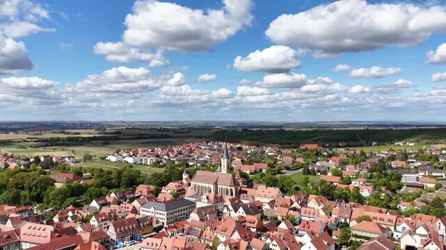 Malerische Panorama-Aussicht auf die historische Altstadt von Iphofen mit dem markanten Stadtturm, umgeben von gr&uuml;nen B&auml;umen unter einem blau-wei&szlig;en Sommerhimmel.