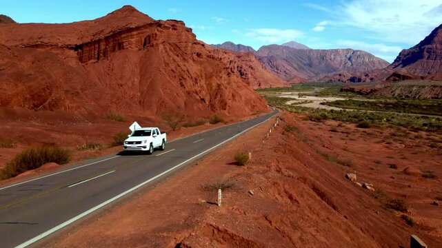 White car travels along a paved highway between red rock cliffs and dry valley in La Quebrada de las Conchas, Salta.
