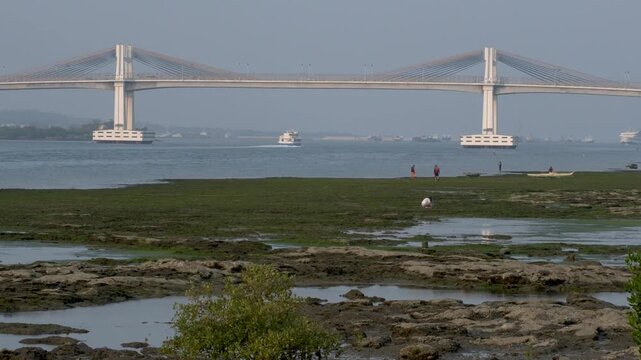 Marcelo Fernan Bridge in Cebu during afternoon golden hour displays a static view of the structure. Low tide exposes the shallow coastal water in the foreground, where people are seen walking along th