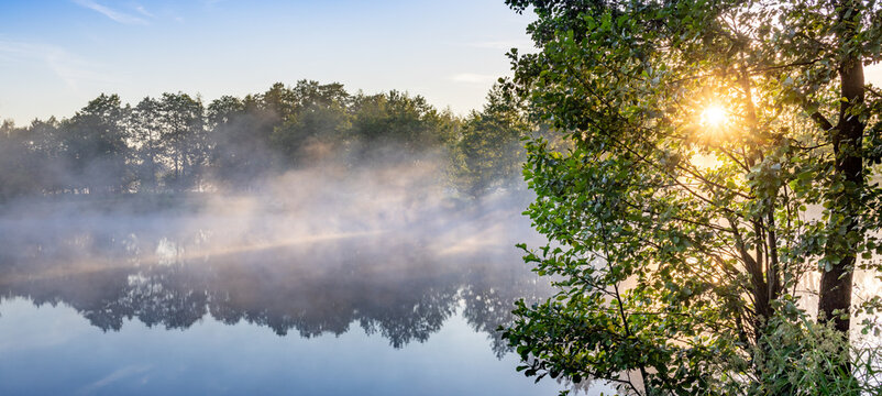 Morning mist hovers over still lake, framed by sunlit trees