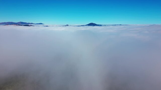 Aerial view above a vast rolling sea of clouds with distant silhouetted mountain peaks breaking through the thick white blanket of morning fog beneath clear sky