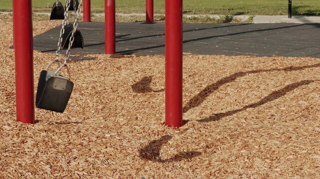 Static shot of an empty playground swing moving slowly over wood chips