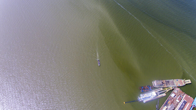 Aerial view of a small boat navigating the waters of Lagos Lagoon near industrial barges and dredging equipment in Oworonshoki, Lagos, Nigeria.