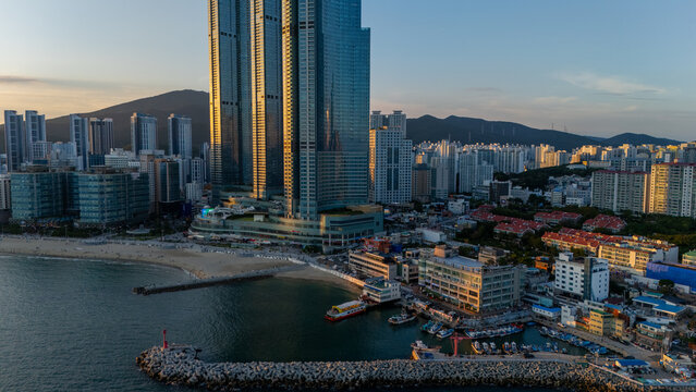 Aerial view of Mipo Port with fishing boats at the breakwater, sandy beach, and towering LCT skyscrapers under the warm glow of the setting sun in Busan, Busan, South Korea.