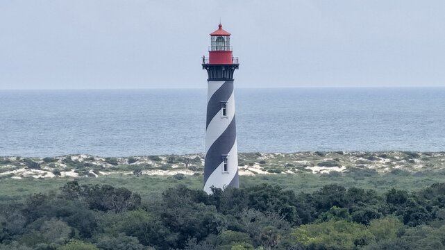 Aerial view of the black and white spiral-striped St. Augustine Lighthouse rising above coastal vegetation and dunes along the Atlantic Ocean in St. Augustine, Florida, United States.
