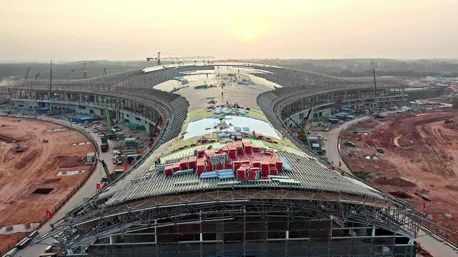 Aerial View of a Large Stadium Under Construction at Sunrise with Steel Roof Framework and Cranes