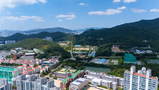 Aerial view of the urban landscape of Haeundae-gu, featuring residential apartment complexes, school buildings, and lush green hills under a clear blue sky in Busan, South Korea.