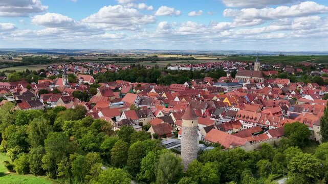 Malerische Panorama-Aussicht auf die historische Altstadt von Iphofen mit dem markanten Stadtturm, umgeben von gr&uuml;nen B&auml;umen unter einem blau-wei&szlig;en Sommerhimmel.