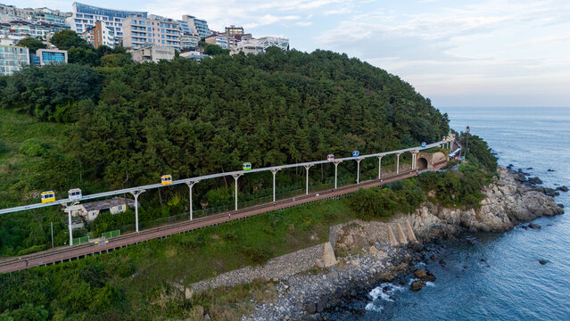 Aerial view of Haeundae Blueline Park with colorful sky capsules moving along the elevated track above the coastal cliffs and lush forest in Busan, South Korea.
