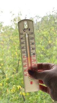 Woman holding a thermometer near a window with yellowing leaves visible outside, representing colder weather, low indoor and outdoor temperatures, autumn and wind.